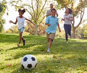 Two children and two adults run on grass toward a soccer ball in a sunlit park with trees in the background.