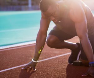 A person in athletic gear is kneeling at the starting line on an outdoor track, preparing to run.